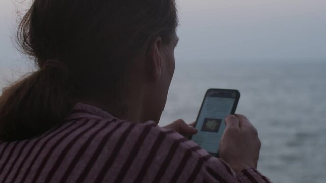Close up over the shoulder view of man scrolling newsfeed on defocused sea background. Faceless man holding smartphone reading breaking news outdoors slow motion copy text space. Information concept