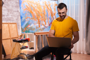 Man painter in studio searching for inspiration on laptop in art studio. Modern artwork paint on...