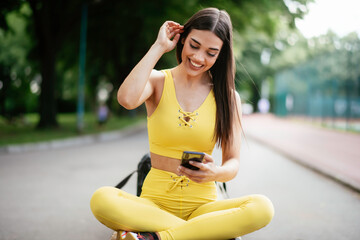 Beautiful woman resting from working out. Young athlete woman sitting on bench and using the phone after training.	