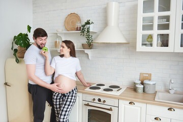 Cute and funny future parents in home outfit embrace standing in the kitchen. Pregnant wife gives husband a green apple. Healthy Lifestyle, parenthood, maternity, expectation of a child, copy space.