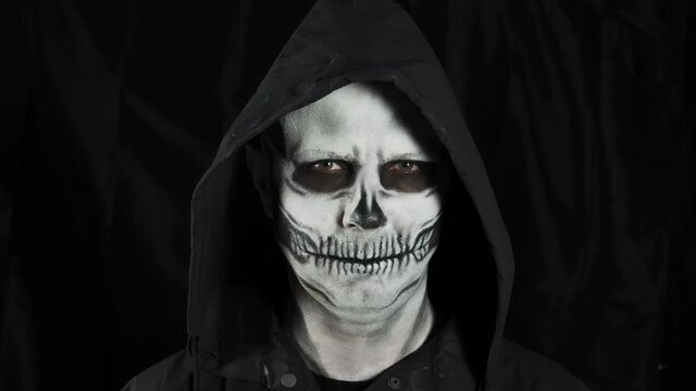 Man with make-up skeleton and black hood on a dark background. A man takes off his hood showing a skull. Halloween or horror theme.