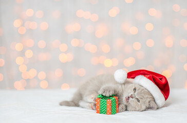 Playful kitten wearing red santa's hat lies with gift box and looks up on empty space. Festive background. Empty space for text