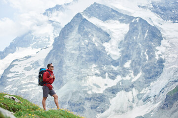 Naklejka premium Horizontal snapshot of a huge snowy mountain in Swiss Alps which takes all the background, male hiker with a backpack standing on green meadow on foreground. Concept of tourism, hiking, trekking.