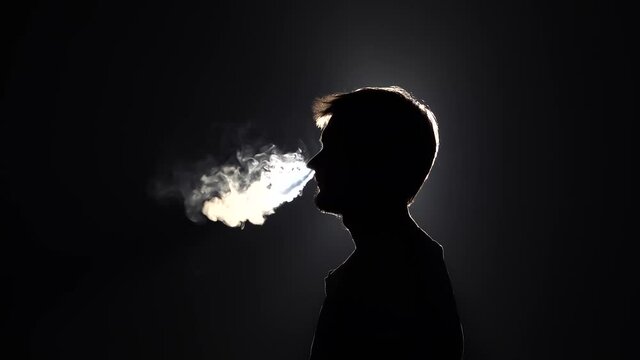 Profile view of silhouette bearded man blowes smoke through the nose in slow motion on black background, close-up