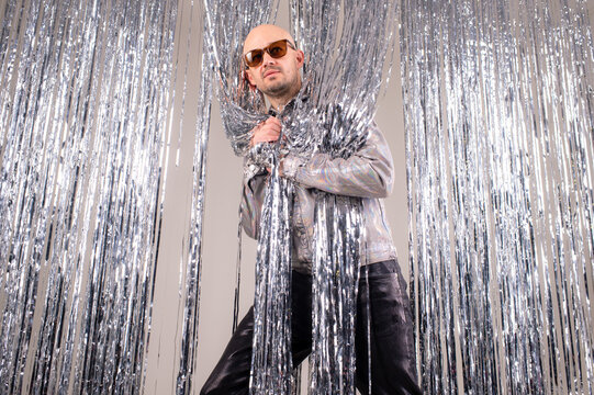 Strange caucasian man in a glittering white shirt, black leather pants, grey shoes and black sunglasses fooling around shiny silver tinsel