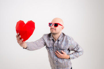 Lovely caucasian man without hair in grey shirt, black leather trousers, red sunglasses and grey trainers with volume red heart
