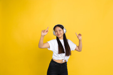 Dancing while listen to music with headphones, pointing. Portrait of young asian woman isolated on yellow studio background. Human emotions, facial expression, sales, ad, shopping concept. Copyspace.