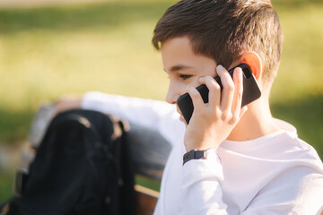 Young boy in white sweatshirt with black backpack sitting on the bench in the park and speak with somebody by the phone