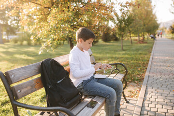 Handsome teenage boy take from his backpack powerbark for charging smarphone. Cute boy in the park in autumn time