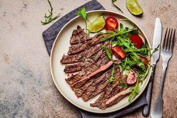 Sliced grilled beef steak with arugula and tomato salad in white plate, dark background, copy space.