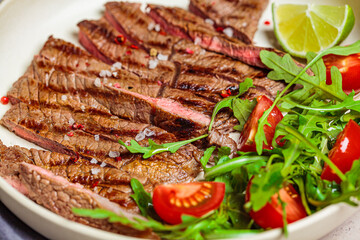 Sliced grilled beef steak with arugula and tomato salad in white plate, dark background, close-up.
