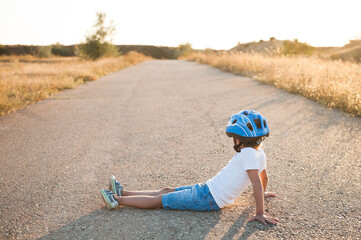 tired small kid wearing sport equipment helmet and sneakers sitting on empty asphalt road on summer warm sunset