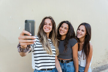 Three young women taking a selfie together on the yellow wall with the smartphone - Beautiful millennials having fun with social network in the city - Copy space