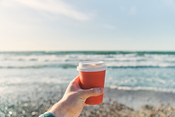 A young man holds a paper cup with coffee on the background of the sea. High-quality photo