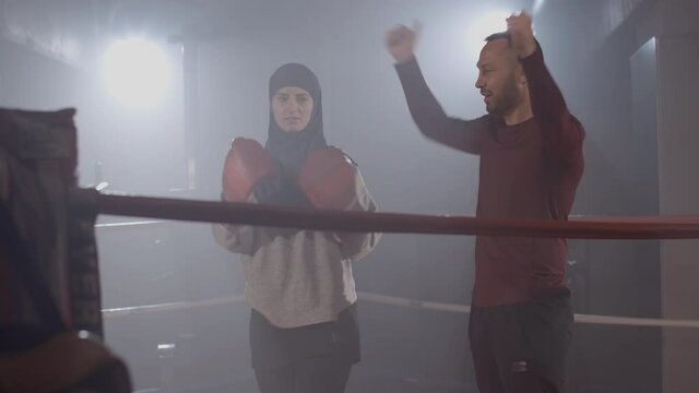 Cheerful Caucasian trainer showing boxing punch to muslim woman in foggy gym with backlight at the background. Portrait of personal instructor training female boxer. Cinema 4k ProRes HQ.