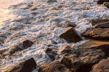 natural filled frame wallpaper background early morning photo of white foamy waves crashing and splashing against sharp brown rocks with the reflection of the sun in the sea