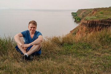 Naklejka na ściany i meble A blond man sits on the grass in front of a rock from the sea