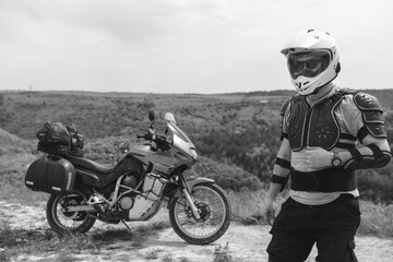 Fototapeta na wymiar Biker man in a helmet and motorcycle equipment, jacket turtle, body armor. Close. In the background a touristic motorcycle. The concept of extreme and hobby. Traveling by bike. Black and white