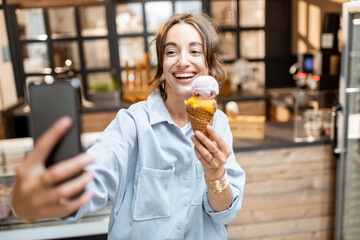 Young and cheerful woman making selfie photo with yummy ice cream in waffle cone, having fun in the...