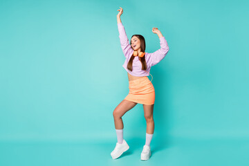 Full size photo of cute millennial lady listen modern technology earphones dance overjoyed raise hands wear cropped violet sweater orange skirt sneakers isolated teal color background