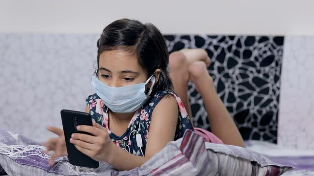 Cute little girl having a video call via smartphone - distance communication concept. Sick Indian child/kid in surgical mask doing video call to her school friends during the Covid-19 pandemic time