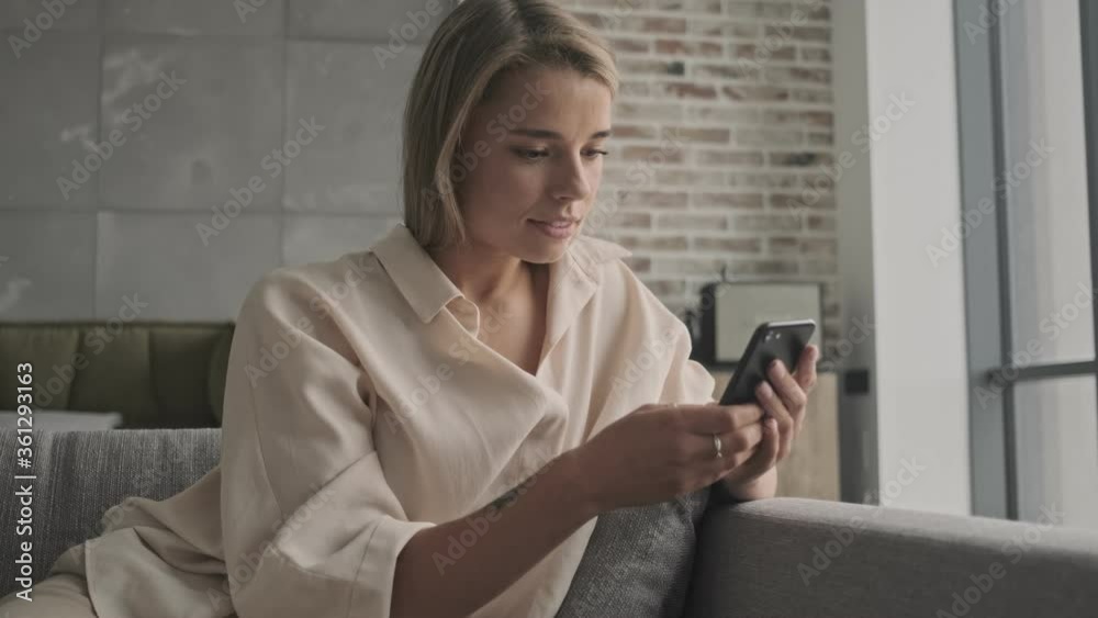 Wall mural Smiling attractive blonde woman using smartphone while sitting on sofa at home