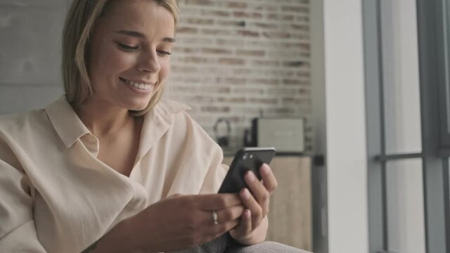 Happy attractive blonde woman using smartphone then looking at window while sitting on sofa at home