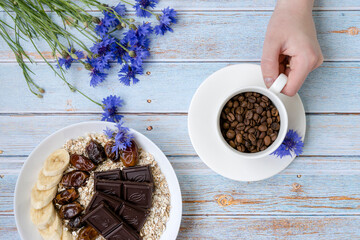 Hand holds a cup with grains of coffee. Oatmeal with bananas and dates in a plate. Cornflowers on a boardwalk blue background. Healthy breakfast. Diet. Ingredients