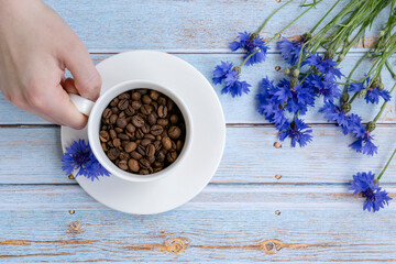 Obraz na płótnie Canvas Hand holds a cup with grains of coffee. Cornflowers on a boardwalk blue background. View from above. White cup of espresso. Ingredients for coffee.
