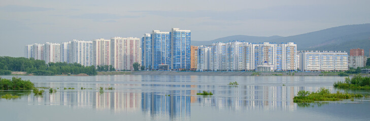 View of the pond. Islands with green vegetation. In distance, typical high-rise buildings, mountains, sky. On surface of water is reflection. Concept of a residential area of the city.
