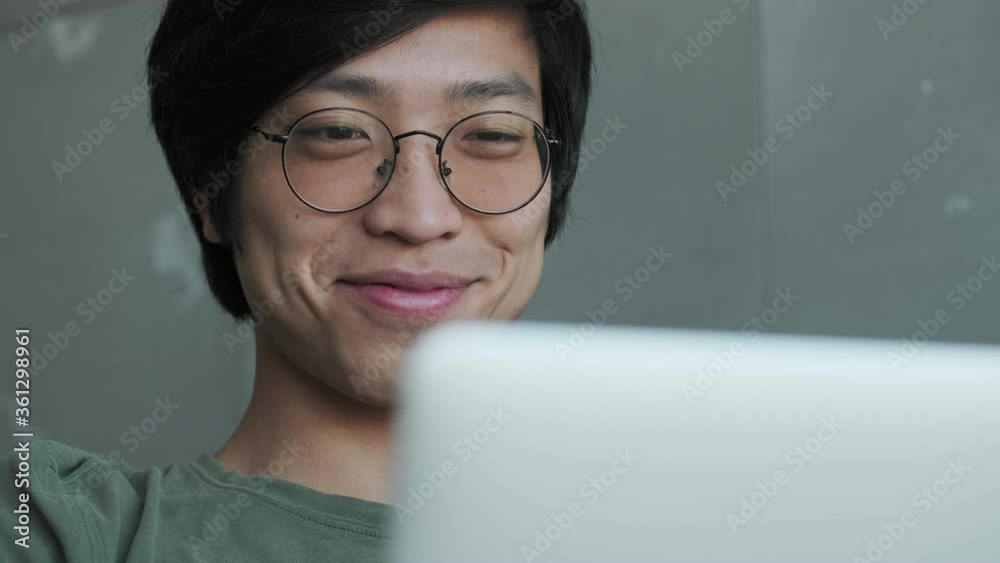 Sticker A close-up view of a happy pleased young asian man wearing eyeglasses is looking to his laptop sitting on couch in apartment