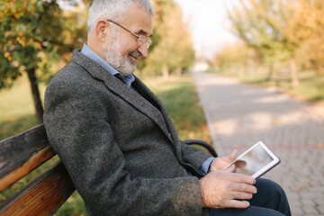 Fototapeta premium Mock up of senior man using tablet outside. Back view of elderly man sitting on the bench and using tablet