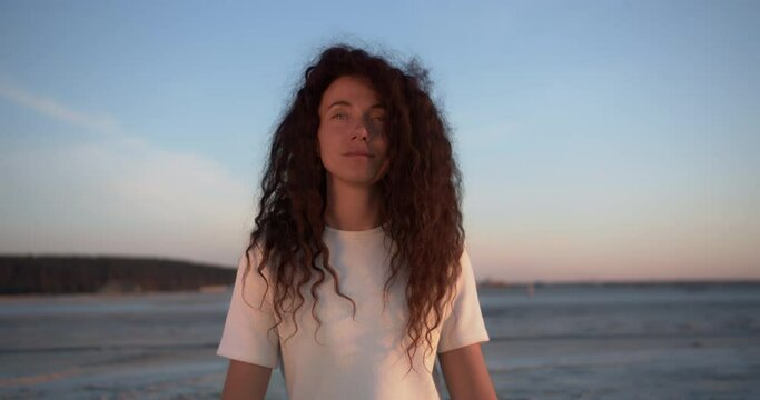 A girl with long curly hair and a natural beautiful face stands against a soft sunset landscape of a sandy beach, she looks at the camera and breathes deep fresh air