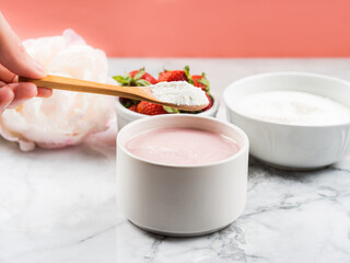 Collagen protein powder in bowl with wooden spoon on marble table. Adding collagen supplement to strawberry yogurt. Female hand