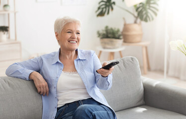 Fototapeta na wymiar Happy elderly lady watching tv at home, sitting on couch with controller