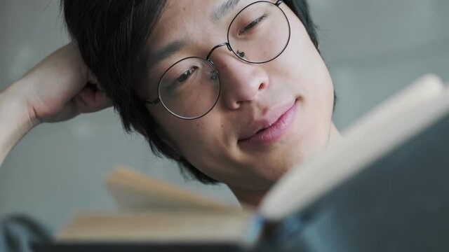A close-up view of a relaxed young asian man wearing eyeglasses is reading a book while sitting in the living room at home