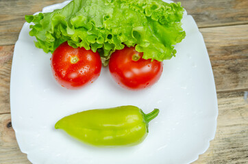Fresh juicy vegetables peppers, tomatoes, cucumbers lettuce leaves with water drops beautifully laid out on a white plate close-up. Vegetables are in the form of a funny face, view top