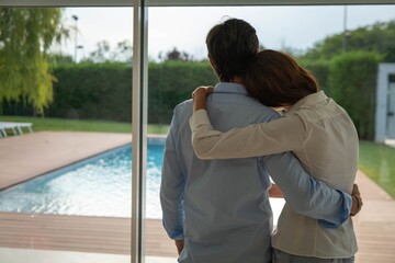 Authentic back view shot of lovely embraced married couple is enjoying time together while looking through a window with a view on a garden with pool at home.