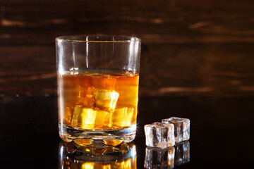 Glass of whiskey with ice cubes on black table against wooden background