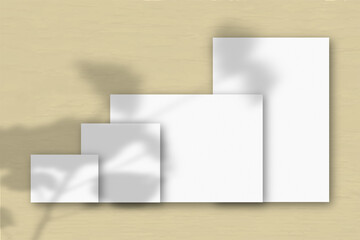 Several horizontal and vertical sheets of white textured paper against a yellow wall background. Mockup with an overlay of plant shadows. Natural light casts shadows from a tropical plant