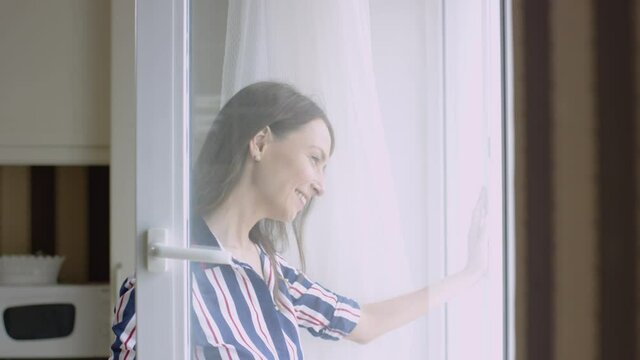 Beautiful woman looks out window happily. Video. Bright woman with smile looks out window watching children. Attractive mother happily watches her child from window