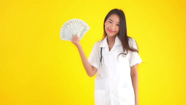Portrait of asian female doctor showing dollar bills. Huge sallary for medical workers concept, static slow motion, isolated on yellow background