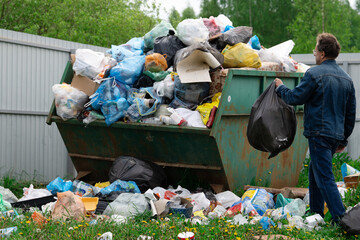 Man in denim suit throws a plastic bag with a garbage to junk container outdoors at countryside