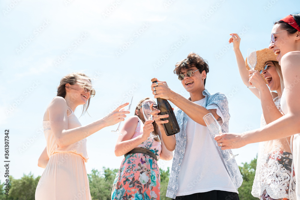 Wall mural Opening champagne, clinking glasses. Seasonal feast at beach. Group of friends celebrating, resting, having fun in sunny summer day. Look happy and cheerful. Festive time, wellness, holiday, party.