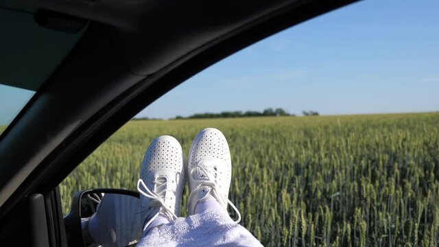 Legs of girl in car window, riding car on country road past wheat field. free woman travels by car. healthy Young woman enjoys traveling by car, protruding her legs from an open window. travel concept