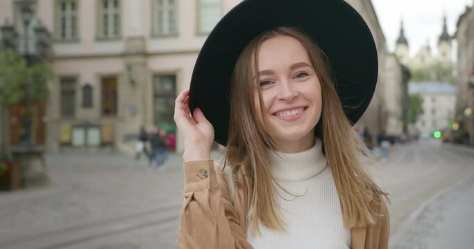 Close-up shooting. The girl is looking up at the camera and smiling. She is holding her hat in her hand. Raindrops are flying past her. The central square of the city in the background. 4K