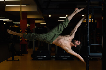 A young man in green pants with a bare torso performs a complex acrobatic element flag on the pylon. professional pole dance in the studio