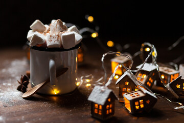 Winter hot drink: hot chocolate with marshmallows, cinnamon and anise. Christmas lights in the shape of houses as decor. Decorative snow, dark wooden background. Festive mood, cozy home atmosphere