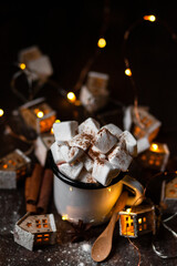 Winter hot drink: hot chocolate with marshmallows, cinnamon and anise. Christmas lights in the shape of houses as decor. Decorative snow, dark wooden background. Festive mood, cozy home atmosphere