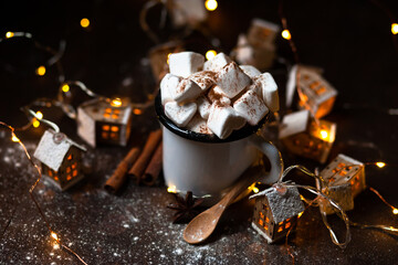 Winter hot drink: hot chocolate with marshmallows, cinnamon and anise. Christmas lights in the shape of houses as decor. Decorative snow, dark wooden background. Festive mood, cozy home atmosphere